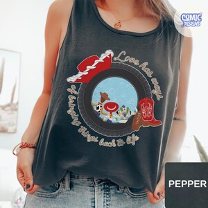 May include: A dark gray sleeveless tank top featuring a graphic print of a tire surrounding a blue sky scene with cartoon characters. The design includes a red cowboy hat, a red cowboy boot, and the text Love has ways of bringing things back to life.