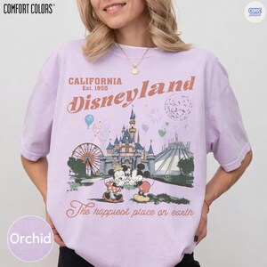 Op de afbeelding: Orchidee-kleurig T-shirt met een Disneyland-afbeelding. Het ontwerp toont het Disneyland-logo, een kasteel, Mickey en Minnie Mouse, en de tekst "The happiest place on earth". Het shirt is van Comfort Colors.