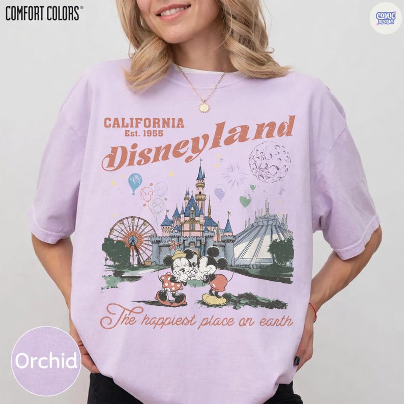 Op de afbeelding: Orchidee-kleurig T-shirt met een Disneyland-afbeelding. Het ontwerp toont het Disneyland-logo, een kasteel, Mickey en Minnie Mouse, en de tekst "The happiest place on earth". Het shirt is van Comfort Colors.