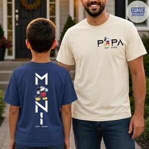 May include: Two t-shirts with Mickey Mouse designs. One is navy blue with a vertical graphic and the text "MZ7" and "EST. 2026". The other is beige with the word "PAPA" and "EST. 2026" with a Mickey Mouse graphic.