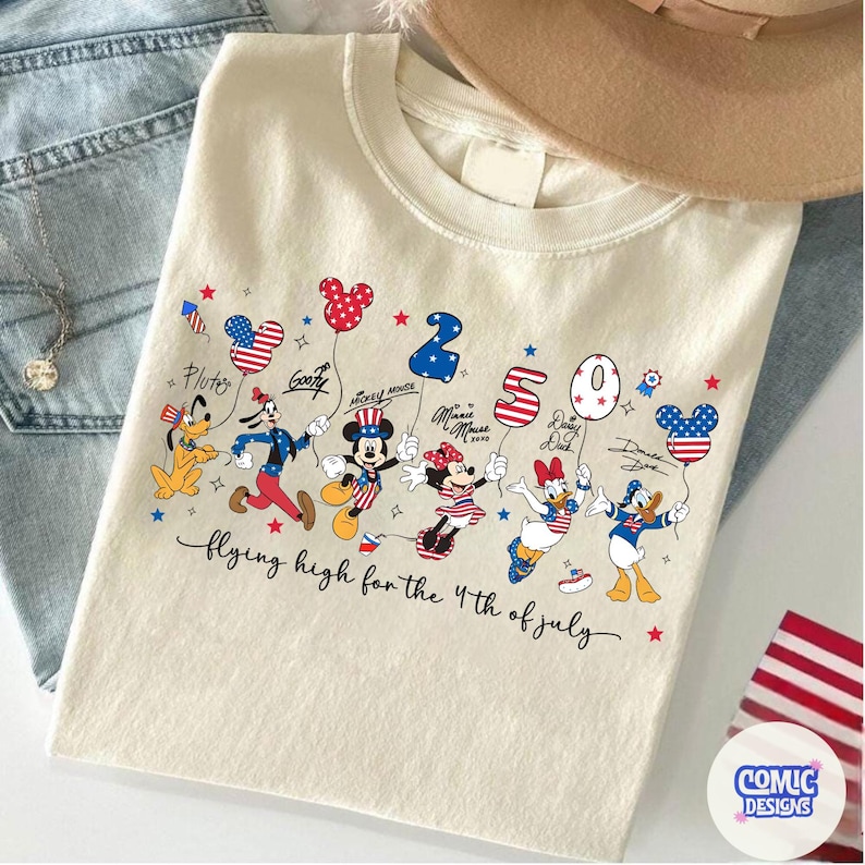 May include: Cream-colored t-shirt featuring Disney characters Pluto, Goofy, Mickey, Minnie, Daisy, and Donald Duck. Each character holds a balloon with an American flag design. The text "2020" and "flying high for the 4th of July" are also visible.