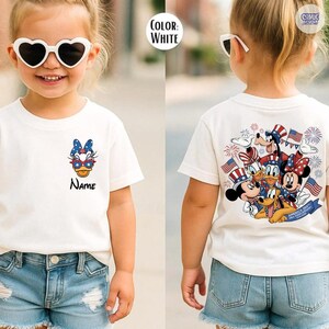 May include: White t-shirt featuring a cartoon character with sunglasses and a bow. The shirt has the word "Name" printed below the character. The back of the shirt features a group of cartoon characters with American flags and fireworks.