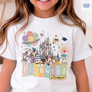 May include: White t-shirt featuring a colorful graphic of Disney characters celebrating graduation. The design includes a castle, balloons, and the words "PreK Grad" and "Congratulations". The name "Elizabeth" is also included.