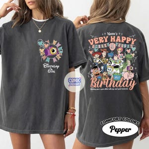 May include: Dark gray t-shirt with a colorful graphic design. The front features a camera and the text "Birthday Girl". The back displays "Very Happy Birthday" with cartoon characters and the text "Hi! Birthday".