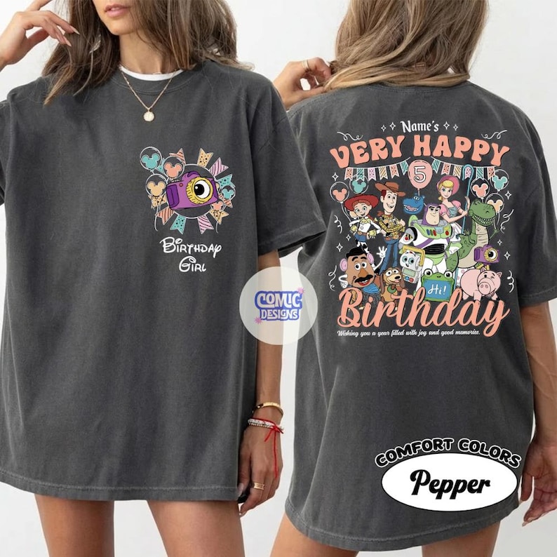 May include: Dark gray t-shirt with a colorful graphic design. The front features a camera and the text "Birthday Girl". The back displays "Very Happy Birthday" with cartoon characters and the text "Hi! Birthday".