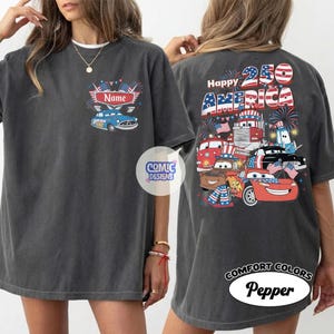 May include: Dark grey t-shirt with a patriotic design. The front has a blue car graphic and the word "Name". The back shows "Happy 250 AMERICA" with cartoon cars, trucks, and fireworks. The shirt is labelled "Comfort Colors Pepper".