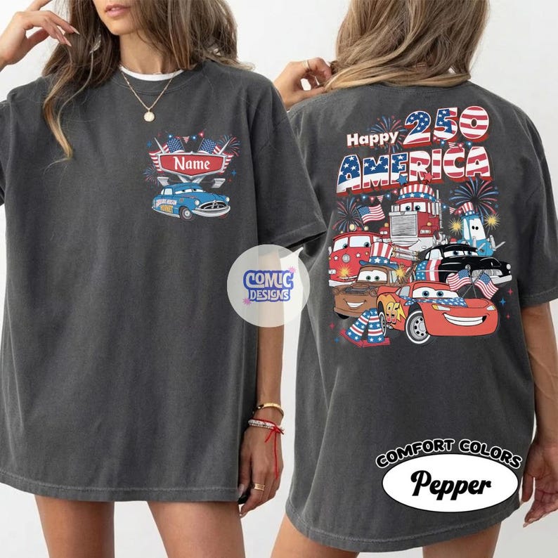 May include: Dark grey t-shirt with a patriotic design. The front has a blue car graphic and the word "Name". The back shows "Happy 250 AMERICA" with cartoon cars, trucks, and fireworks. The shirt is labelled "Comfort Colors Pepper".
