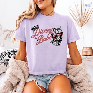 May include: A lilac t-shirt featuring the words "Disney Babe" in a script font, with illustrations of cherries, dice, and a black eight ball. The top has short sleeves and a loose fit.
