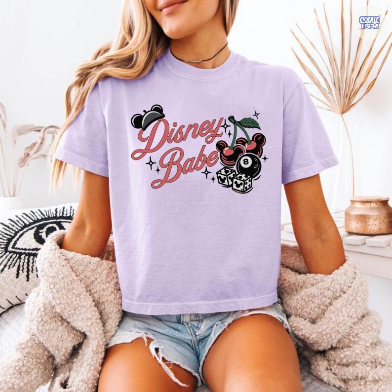 May include: A lilac t-shirt featuring the words "Disney Babe" in a script font, with illustrations of cherries, dice, and a black eight ball. The top has short sleeves and a loose fit.