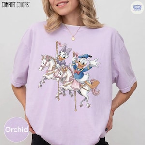 May include: Orchid-colored t-shirt featuring a carousel design with Donald Duck and Daisy Duck riding white horses. The shirt has the words "COMFORT COLORS" at the top left and "COMIC DESIGNS" at the top right. The word "Orchid" is at the bottom left.