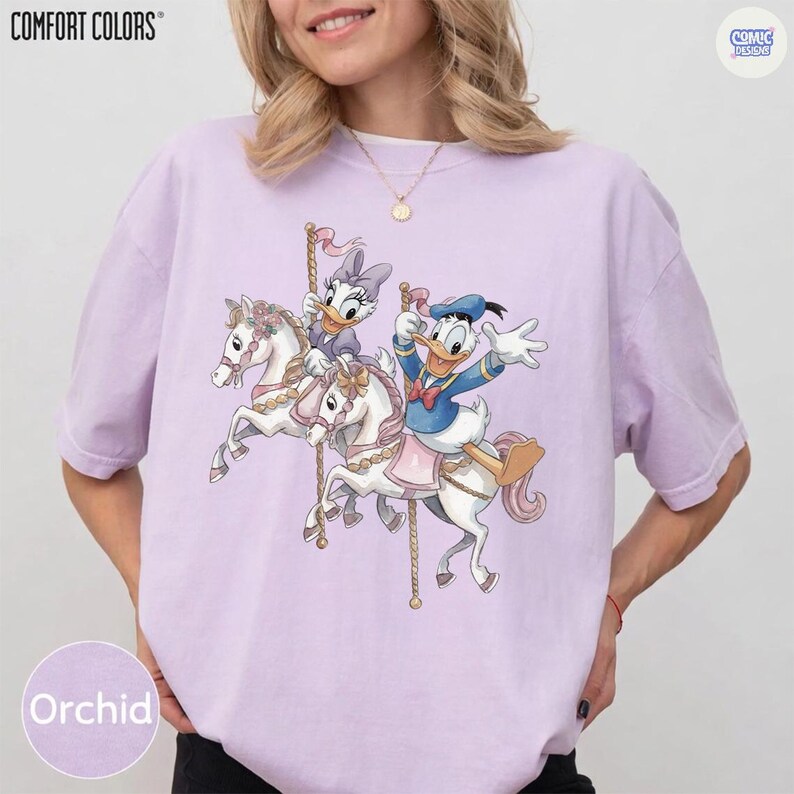 May include: Orchid-colored t-shirt featuring a carousel design with Donald Duck and Daisy Duck riding white horses. The shirt has the words "COMFORT COLORS" at the top left and "COMIC DESIGNS" at the top right. The word "Orchid" is at the bottom left.