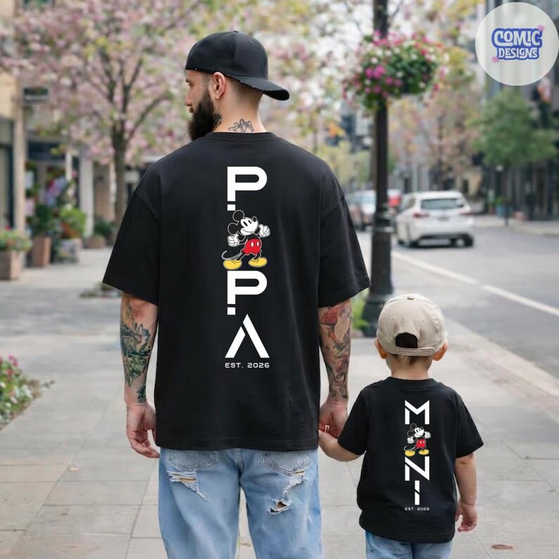 May include: Black t-shirts with a Mickey Mouse graphic and the word "PAPA" in white. The adult shirt has "EST. 2026" printed below the graphic. A child wears a matching shirt with "MINI" and "EST. 2020".