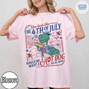 May include: Light pink t-shirt with a graphic featuring a dinosaur in a suit holding a hot dog, with text that reads "You look like the 4th of July, Makes me want a Hot Dog real bad."