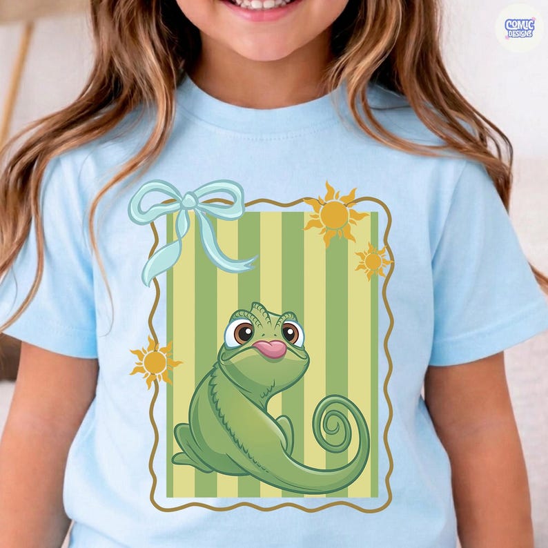 May include: A light blue t-shirt featuring a cartoon chameleon with its tongue out, set against a green and yellow striped background. The design includes a light blue bow and sun graphics. The shirt has the text "COMIC DESIGNS" in the upper right corner.