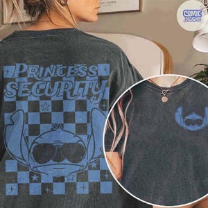 May include: Dark gray t-shirt featuring a blue graphic of a cartoon character wearing sunglasses, with the text "PRINCESS SECURITY" on the back. A smaller version of the character is on the front. The design includes a checkered pattern.
