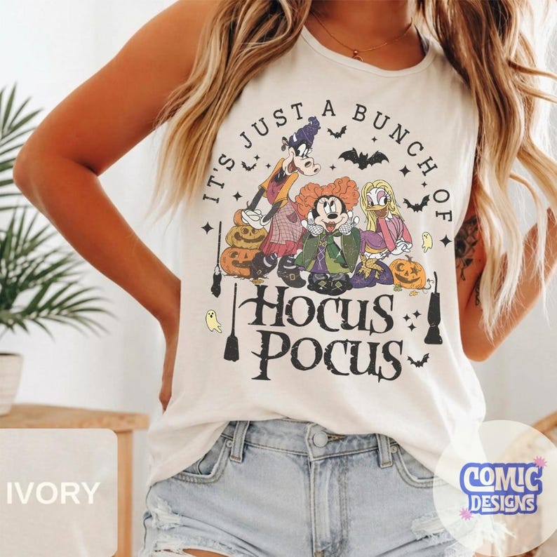 May include: An ivory sleeveless tank top featuring a graphic print of three cartoon characters dressed as witches, surrounded by pumpkins, bats, and ghosts. The text reads, "IT'S JUST A BUNCH OF HOCUS POCUS" in a black, stylized, serif font.