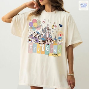 May include: Cream-colored t-shirt featuring a colorful graphic of Disney characters, a castle, balloons, and the words "Prek Grad" and "Congratulations." The design includes Mickey Mouse, Minnie Mouse, Donald Duck, and Goofy. The shirt has a relaxed fit.