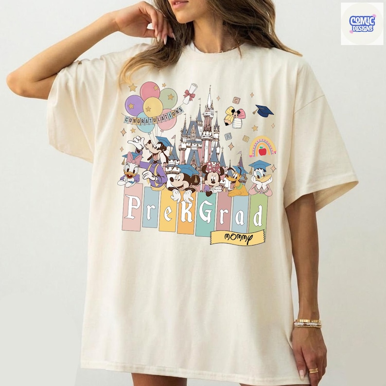 May include: Cream-colored t-shirt featuring a colorful graphic of Disney characters, a castle, balloons, and the words "Prek Grad" and "Congratulations." The design includes Mickey Mouse, Minnie Mouse, Donald Duck, and Goofy. The shirt has a relaxed fit.