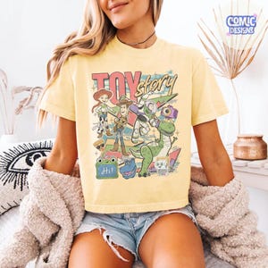 May include: A pale yellow short-sleeve t-shirt featuring a colorful graphic print of Toy Story characters including Woody, Jessie, Buzz Lightyear, Rex, and others. The design includes the text Toy Story and a small green frog with the word Hi.