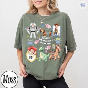May include: An olive green short-sleeve t-shirt featuring a graphic print of various animated characters including a space ranger, a cowboy, a cowgirl, a dinosaur, a spring dog, and a horse. The text "YOU'VE GOT A FRIEND IN ME" is printed in the center.