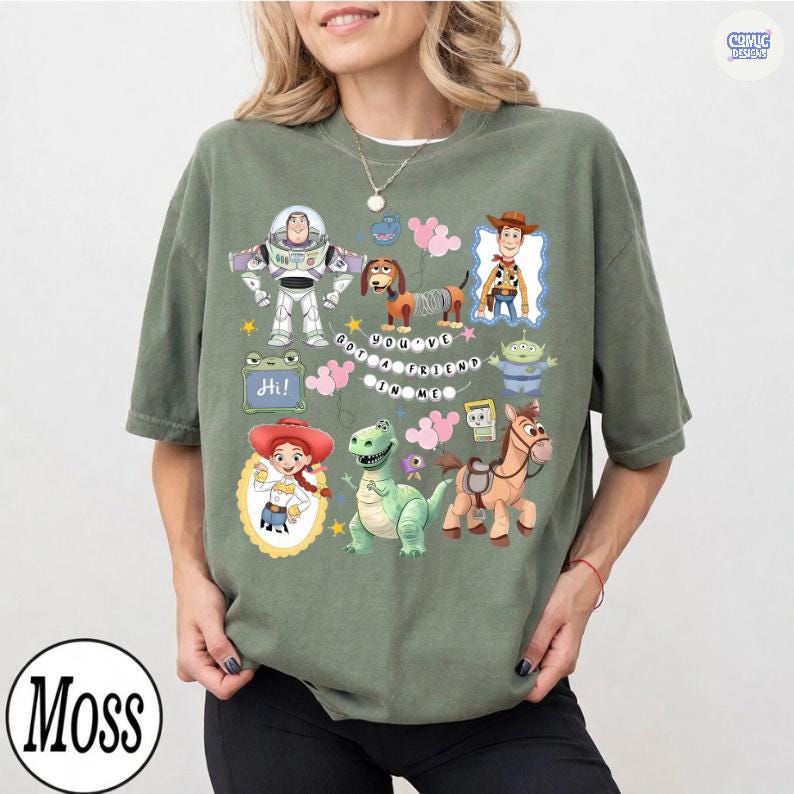 May include: An olive green short-sleeve t-shirt featuring a graphic print of various animated characters including a space ranger, a cowboy, a cowgirl, a dinosaur, a spring dog, and a horse. The text "YOU'VE GOT A FRIEND IN ME" is printed in the center.