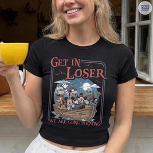 May include: Black short-sleeve t-shirt with a pirate-themed graphic. The graphic features cartoon characters in a boat with the text "GET IN LOSER" above and "WE ARE GOING PIRATING" below. A "Comic Designs" logo is in the upper right corner.