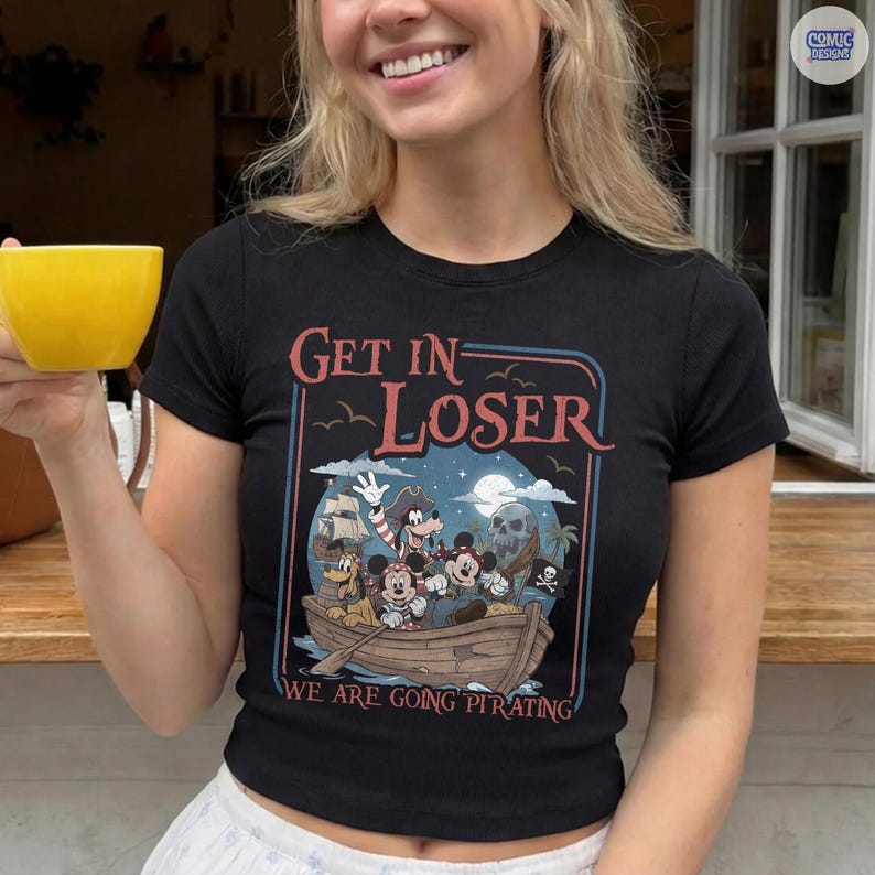 May include: Black short-sleeve t-shirt with a pirate-themed graphic. The graphic features cartoon characters in a boat with the text "GET IN LOSER" above and "WE ARE GOING PIRATING" below. A "Comic Designs" logo is in the upper right corner.