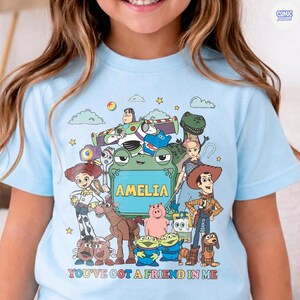 May include: A light blue t-shirt featuring a colorful graphic of various animated characters from the Toy Story franchise surrounding a central green frog-shaped frame with the name AMELIA printed inside and the text YOU'VE GOT A FRIEND IN ME.