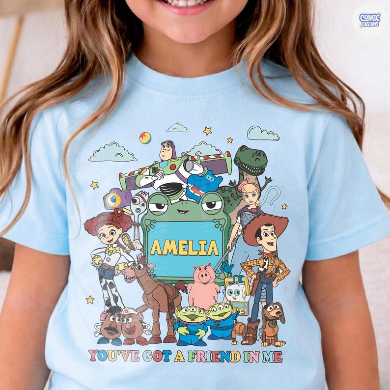 May include: A light blue t-shirt featuring a colorful graphic of various animated characters from the Toy Story franchise surrounding a central green frog-shaped frame with the name AMELIA printed inside and the text YOU'VE GOT A FRIEND IN ME.