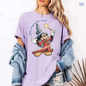 May include: A lilac t-shirt with a graphic of Mickey Mouse in a sorcerer's outfit, holding a wand with a starburst. The text "some imagination huh?" is printed on the shirt. The sleeves are rolled up.