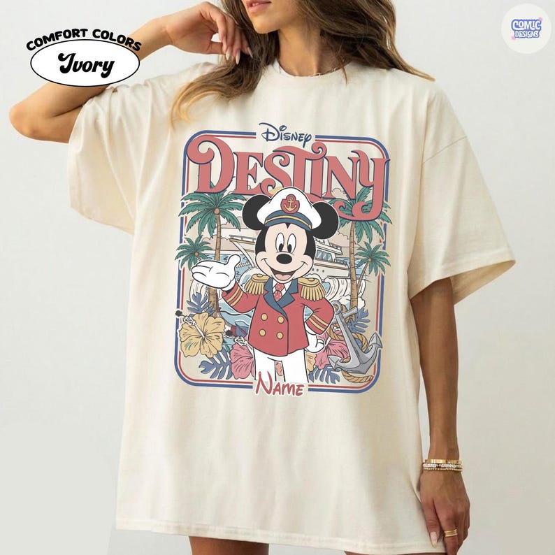 Op de afbeelding: Ivoor Comfort Colors T-shirt met een Disney-ontwerp. De afbeelding toont Mickey Mouse in een kapiteelsuniform, het woord "Destiny" en tropische elementen. Het shirt heeft korte mouwen en een ronde hals.