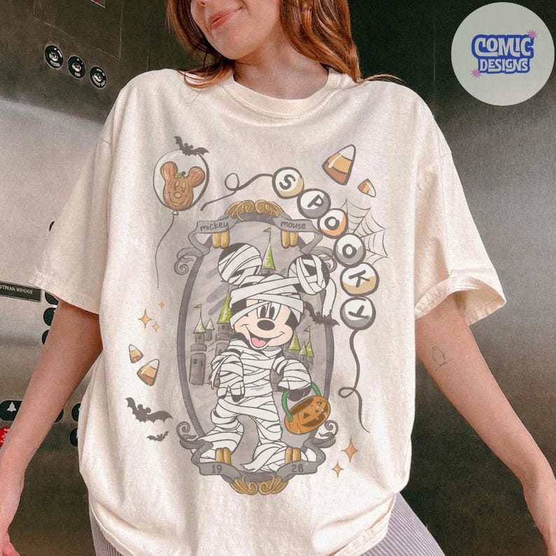 Puede incluir: Una camiseta de color crema con un gr&aacute;fico de un rat&oacute;n de dibujos animados envuelto en vendas como una momia, sosteniendo un cubo de calabaza. El dise&ntilde;o incluye un castillo, murci&eacute;lagos, ma&iacute;z dulce, telara&ntilde;as y el texto SPOOKY y mickey mouse 1928.