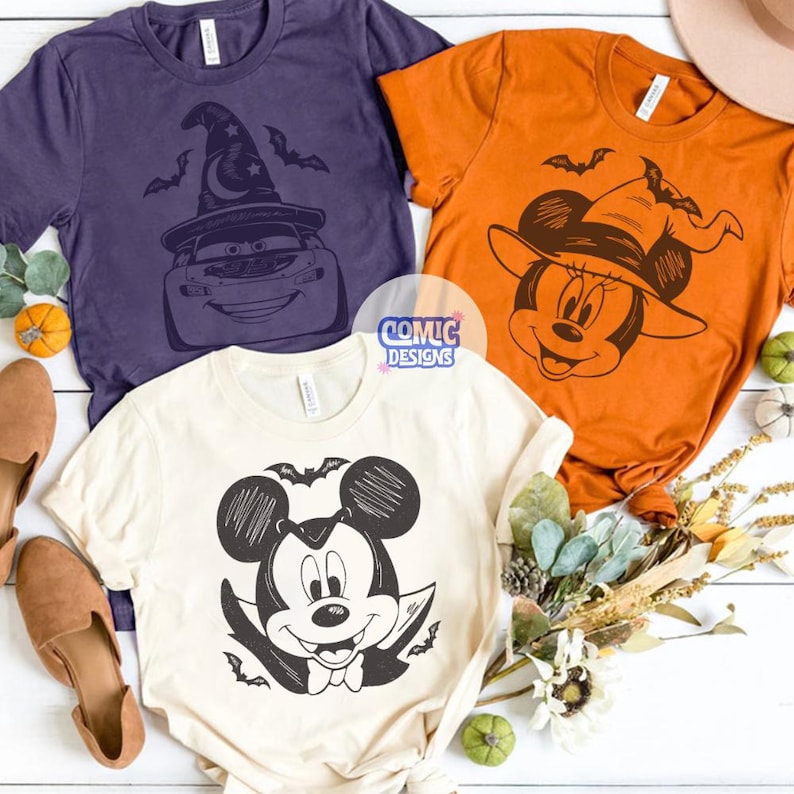 May include: Three Halloween-themed t-shirts in purple, orange, and cream. The purple shirt features a cartoon car wearing a witch hat. The orange shirt has a cartoon mouse in a witch hat. The cream shirt displays a cartoon mouse in a vampire costume. The shirts have the text "COMIC DESIGNS".