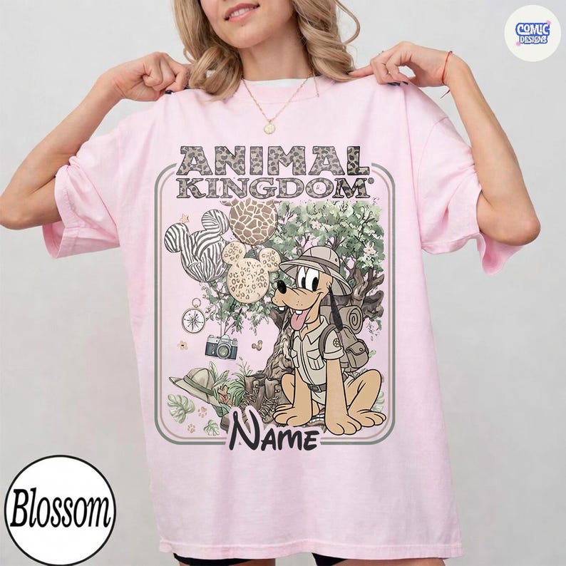 May include: A light pink t-shirt featuring a cartoon illustration of Pluto in safari attire, with the text "ANIMAL KINGDOM" above and "NAME" below. The design includes Mickey Mouse head balloons and a tree.