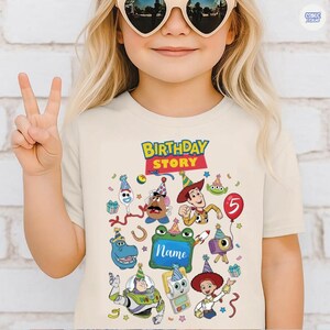 May include: A cream-colored t-shirt featuring a colorful graphic print with the text "BIRTHDAY STORY" and "Name" in a central frame. The design includes various cartoon characters wearing party hats, a red balloon with the number 5, and confetti.