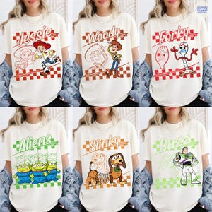 May include: A collection of six off-white t-shirts featuring Toy Story characters. Each shirt displays a character name, a line drawing, and a full-color illustration of Jessie, Woody, Forky, Aliens, Slinky, or Buzz Lightyear over a checkered pattern.