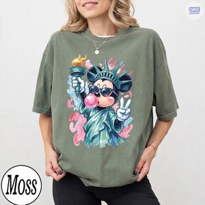 May include: An olive green t-shirt featuring a graphic of a cartoon mouse dressed as the Statue of Liberty, wearing sunglasses, holding a torch, and blowing a pink bubble. The design includes abstract pink and blue paint splatters in the background.