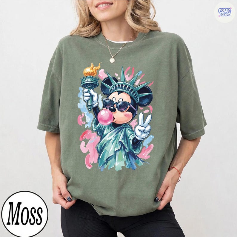 May include: An olive green t-shirt featuring a graphic of a cartoon mouse dressed as the Statue of Liberty, wearing sunglasses, holding a torch, and blowing a pink bubble. The design includes abstract pink and blue paint splatters in the background.