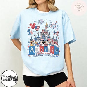 May include: A light blue t-shirt featuring a graphic of cartoon characters in front of a castle with fireworks. The design includes the text America, 250 Years of Freedom, and 1776-2026 in red, white, and blue colors with star and bow accents.