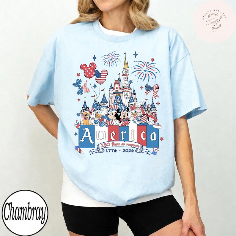 May include: A light blue t-shirt featuring a graphic of cartoon characters in front of a castle with fireworks. The design includes the text America, 250 Years of Freedom, and 1776-2026 in red, white, and blue colors with star and bow accents.
