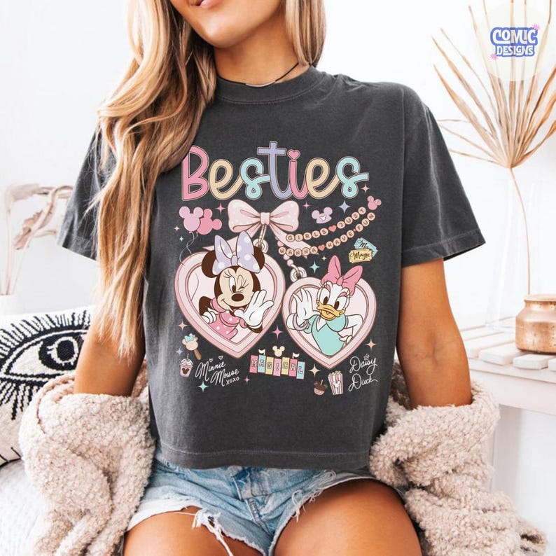 May include: Dark gray t-shirt with the word "Besties" in pastel colors. Features Minnie Mouse and Daisy Duck in heart-shaped frames. Includes the text "Minnie Mouse 2020" and "Daisy Duck".