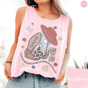 Puede incluir: Una camiseta sin mangas de color rosa claro con un estampado gr&aacute;fico de una bota vaquera con estampado de vaca, un sombrero marr&oacute;n, un lazo, flores y bloques apilados con las letras T, S y la palabra NAME. Un collar de cuentas dice I KNEW IT Y I KNEW IT Y.