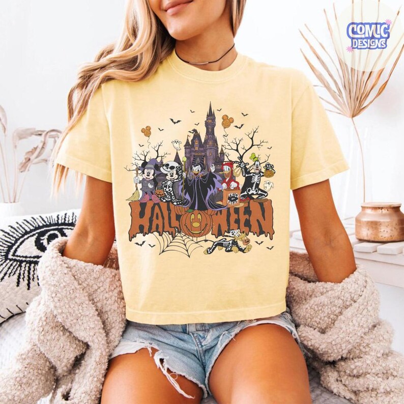 Puede incluir: Una camiseta de color amarillo p&aacute;lido con un estampado gr&aacute;fico de personajes de dibujos animados con disfraces de Halloween frente a un castillo oscuro. El dise&ntilde;o incluye murci&eacute;lagos, una telara&ntilde;a y la palabra HALLOWEEN en letras grandes.