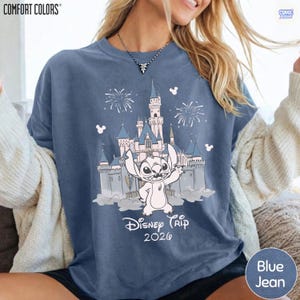 May include: Blue jean Comfort Colors t-shirt featuring a graphic of Stitch in front of a castle with fireworks and the text "Disney Trip 2020". The shirt has a crew neck and short sleeves.
