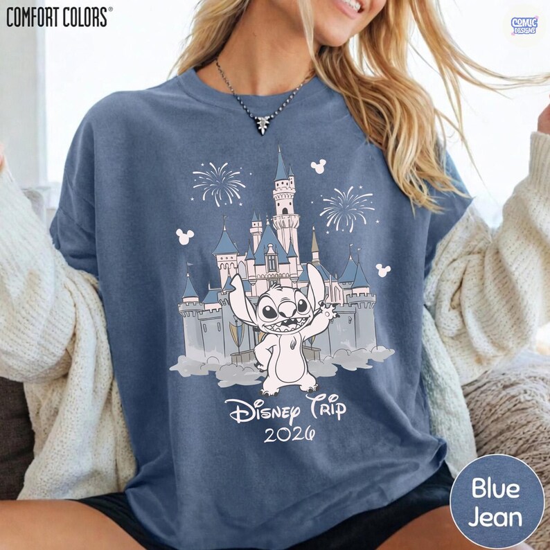 May include: Blue jean Comfort Colors t-shirt featuring a graphic of Stitch in front of a castle with fireworks and the text "Disney Trip 2020". The shirt has a crew neck and short sleeves.