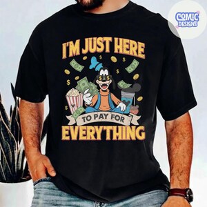 May include: Black t-shirt with a cartoon graphic of Goofy holding money and popcorn, with the text "I'M JUST HERE TO PAY FOR EVERYTHING". The design includes dollar signs and a blue bow tie.