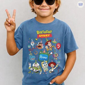 May include: A blue short-sleeved t-shirt featuring a colorful graphic print with the text Birthday Story. The design includes various cartoon characters wearing party hats, a red balloon with the number 5, and a green frame with the word Name.