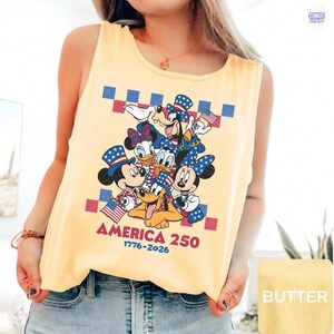 May include: A pale yellow sleeveless tank top featuring a graphic print of Mickey Mouse, Minnie Mouse, Donald Duck, Daisy Duck, Goofy, and Pluto wearing patriotic hats. The text reads AMERICA 250 1776-2026 with a red and blue checkered border.