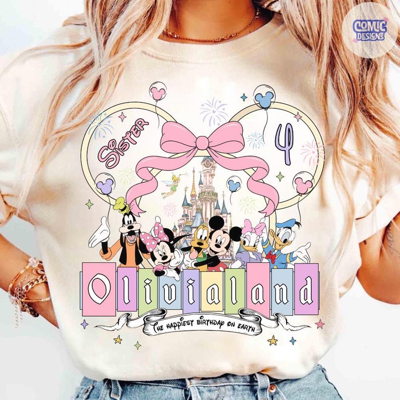 Op de afbeelding: Cr&egrave;mekleurig T-shirt met een kleurrijke Disney-afbeelding. Het toont personages, een roze strik en de tekst "OliviaLand". Het ontwerp bevat het nummer 4, vuurwerk en de zin "The happiest birthday on Earth".