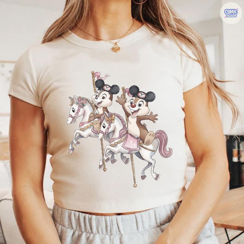 May include: Cream-colored crop top featuring a carousel design with cartoon characters. The carousel horses are white with pink accents, and the characters are brown and black. The top has a crew neck and short sleeves.